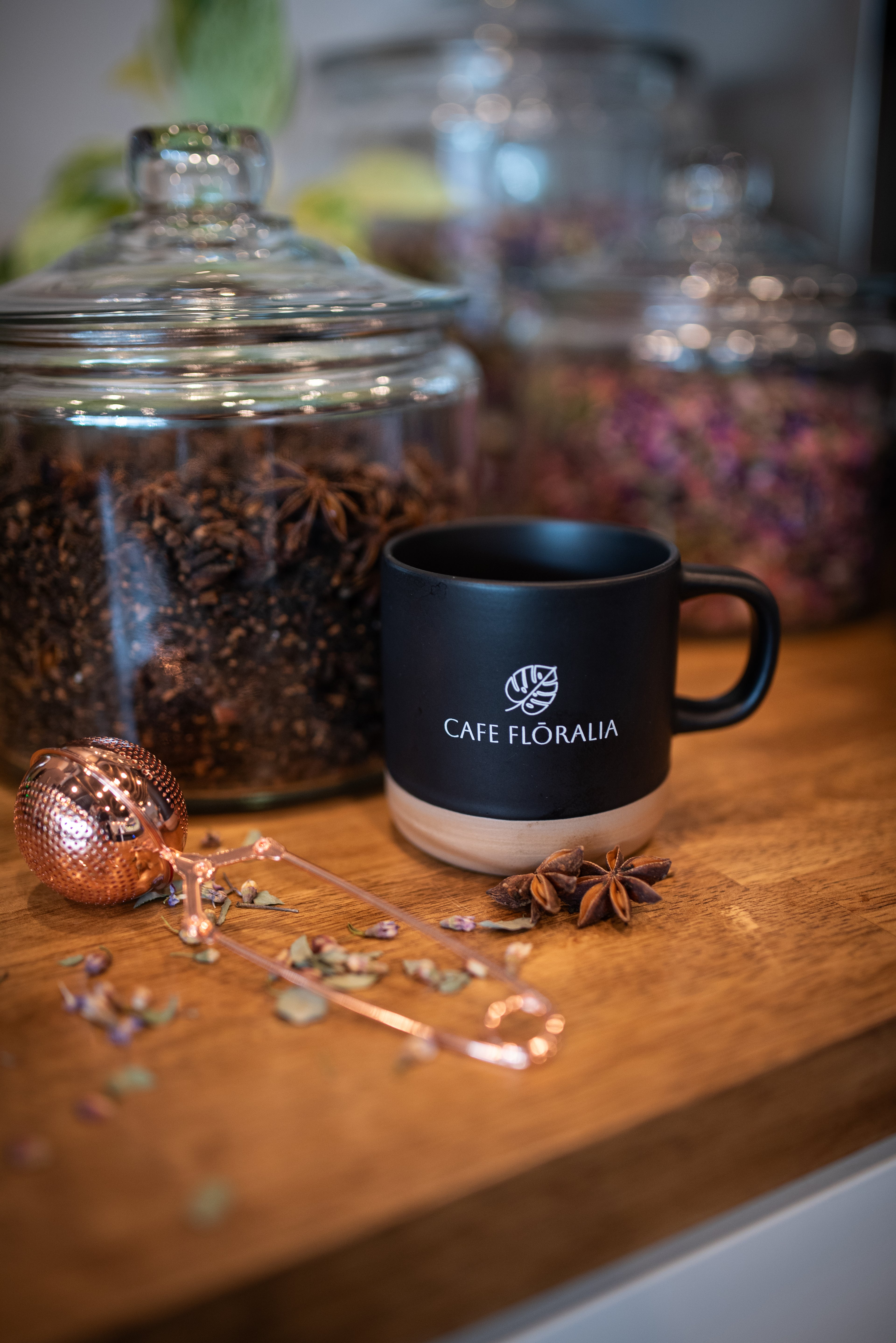 Black mug with 'Cafe Floraia' branding on a wooden surface with jars and herbs.